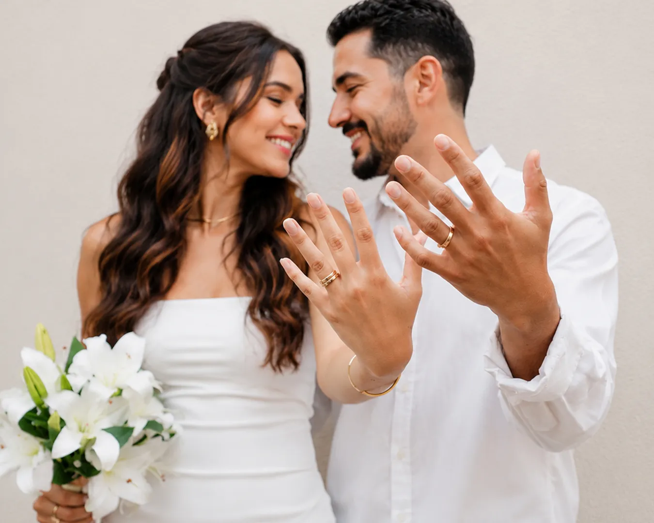 Casal de noivos sorrindo e mostrando as alianças nas mãos, com um buquê de lírios brancos